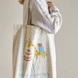 Celebrate Cotton Tote Bag in beige, shown carried over the shoulder, printed with a giraffe in a party hat beside a cake and gifts, surrounded by confetti flecks.