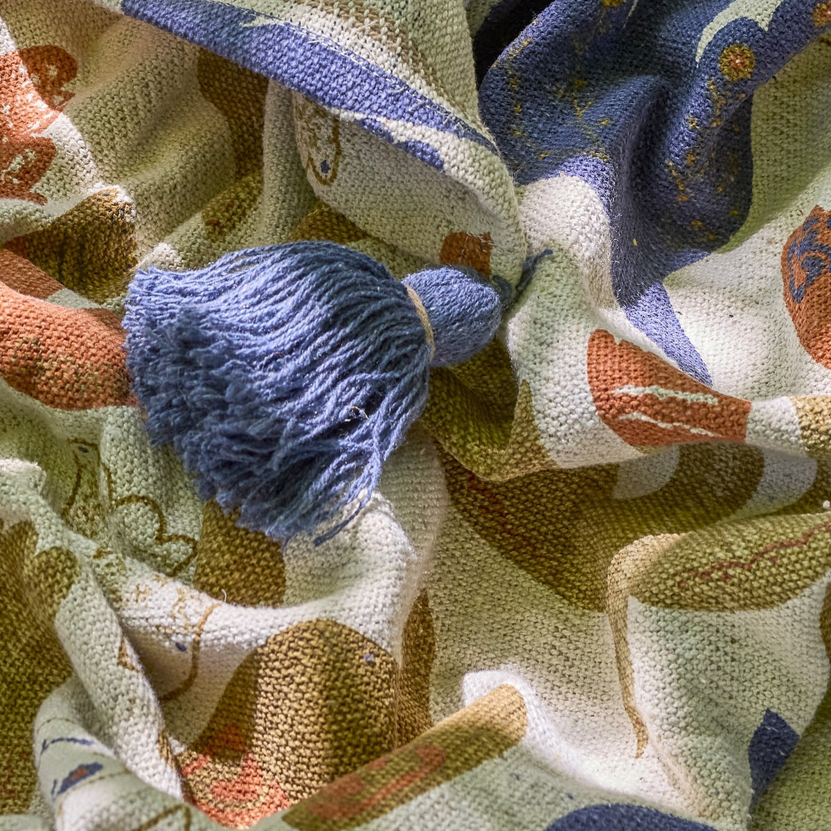 Anniversary Cotton Throw in multicoloured woven cotton, shown rumpled with a blue corner tassel and printed bird and wing pattern in blue, orange, and green.