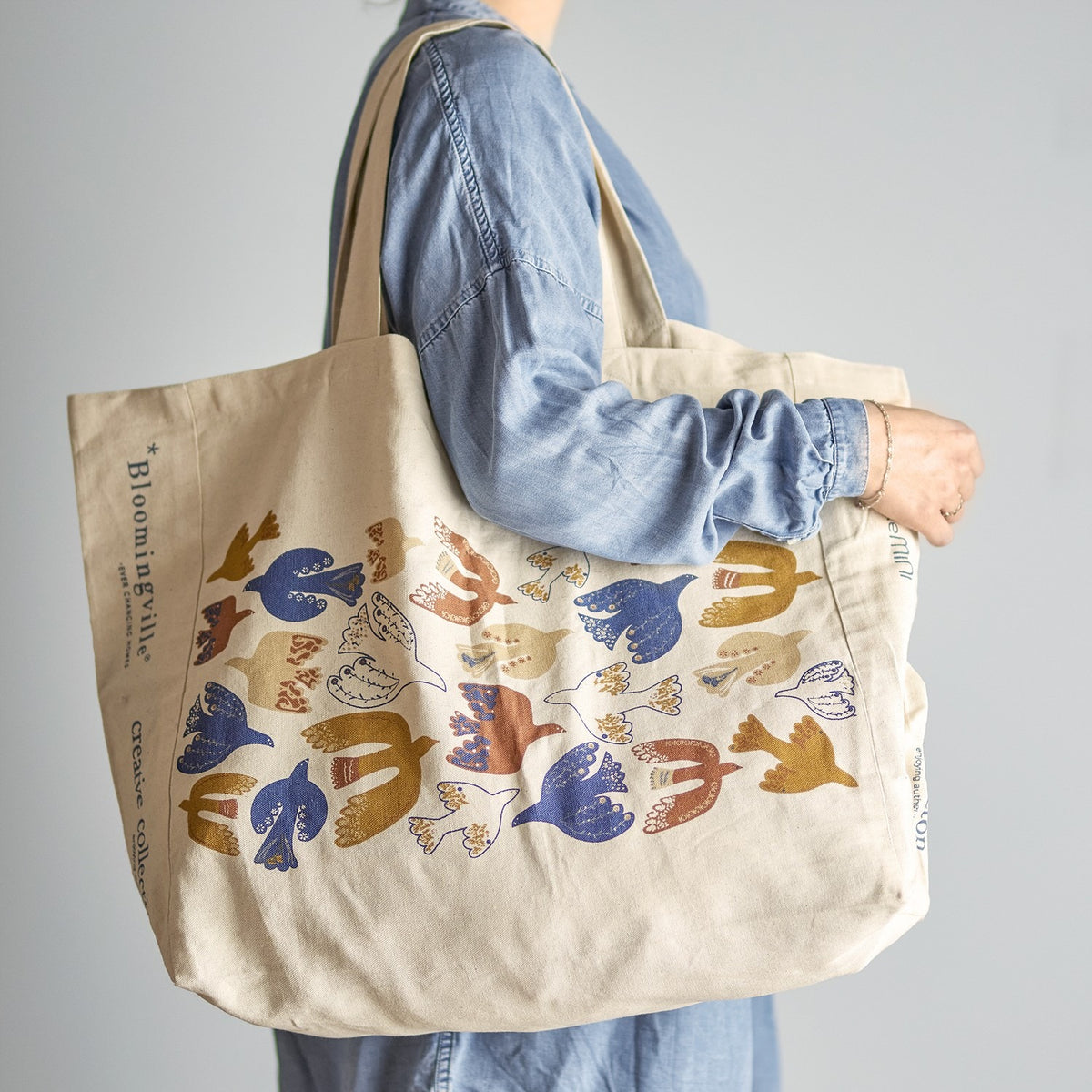 Beige woven cotton shopping bag carried over the shoulder, printed with stylised birds in blue, mustard, rust and cream, with lettering along the side panel.