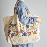 Beige woven cotton shopping bag carried over the shoulder, printed with stylised birds in blue, mustard, rust and cream, with lettering along the side panel.