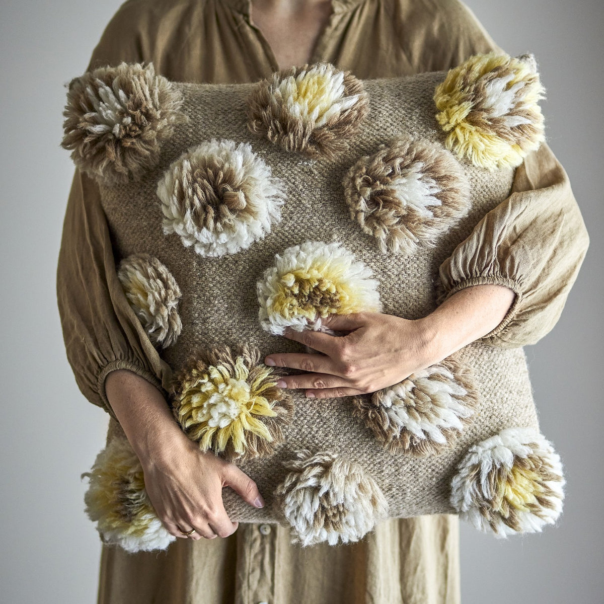 Somma Wool Cushion in Natural, a square cushion with thick, shaggy tufted rosettes in cream, beige and yellow tones, held by a person in a neutral setting.