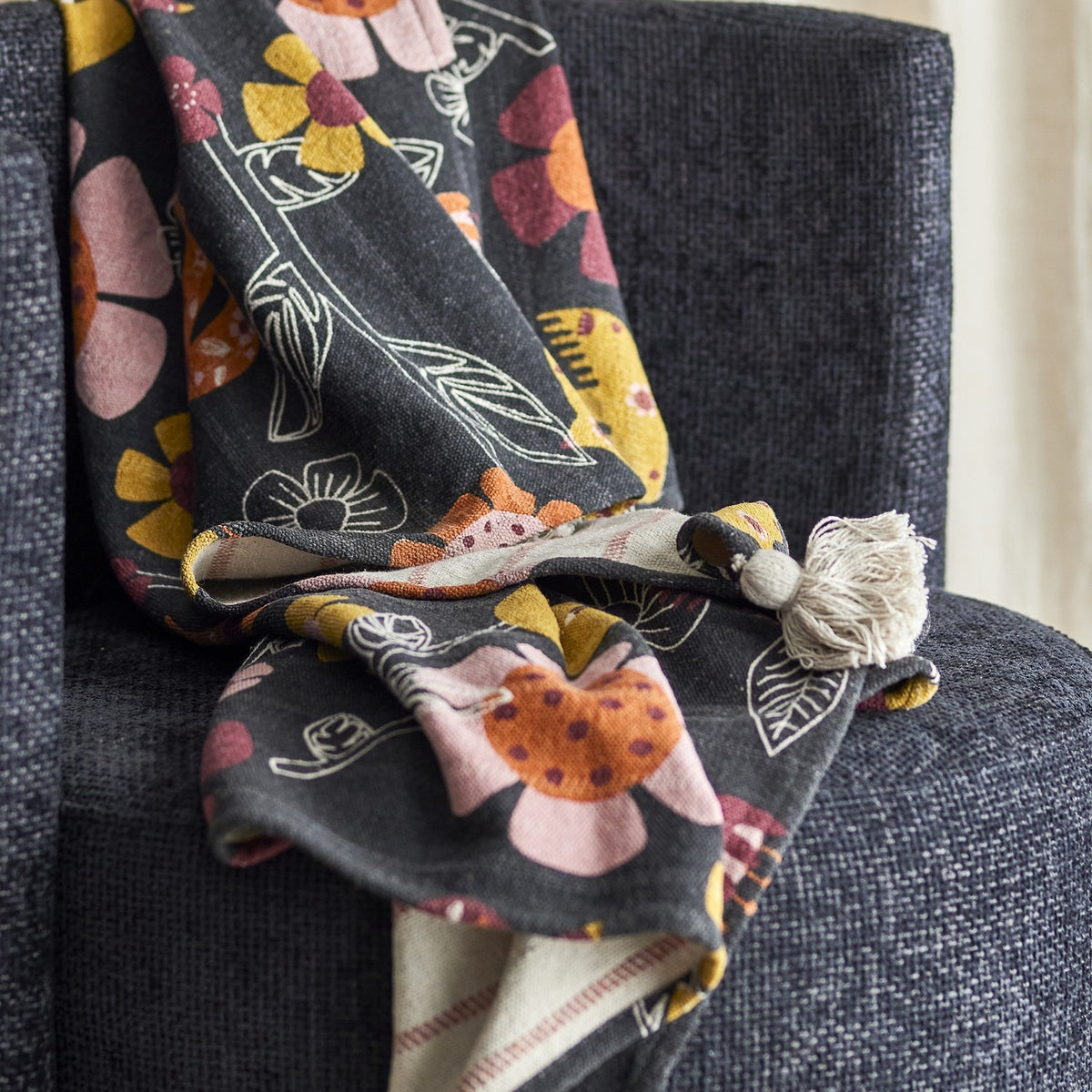 Vivielle Striped Throw draped over a dark grey sofa, showing its floral print face with outlined leaves and birds, tasselled corner, and striped reverse peeking through the folds.