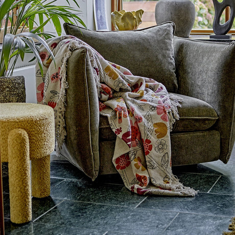 Vivielle Floral Throw in multicoloured cotton-blend fabric with fringed edges, draped over a grey upholstered armchair in a living room setting.