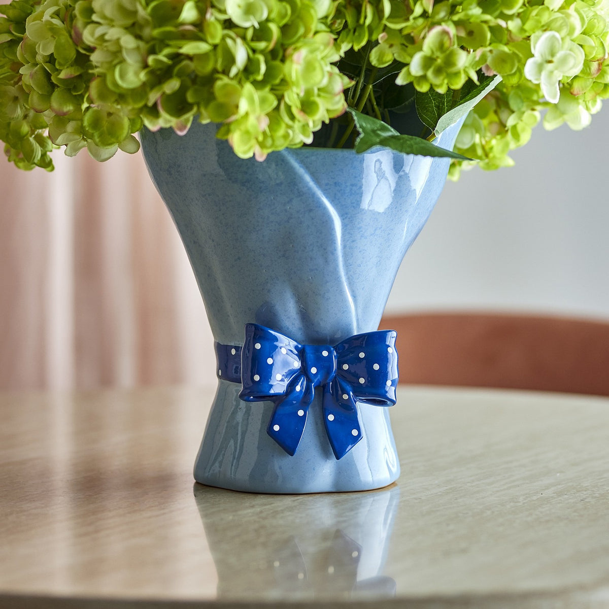Belivia Bouquet Stoneware Vase 28.5cm in speckled blue, shaped like wrapped flowers with a glossy polka-dot bow at the base, shown on a table holding green hydrangeas.