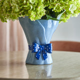 Belivia Bouquet Stoneware Vase 28.5cm in speckled blue, shaped like wrapped flowers with a glossy polka-dot bow at the base, shown on a table holding green hydrangeas.