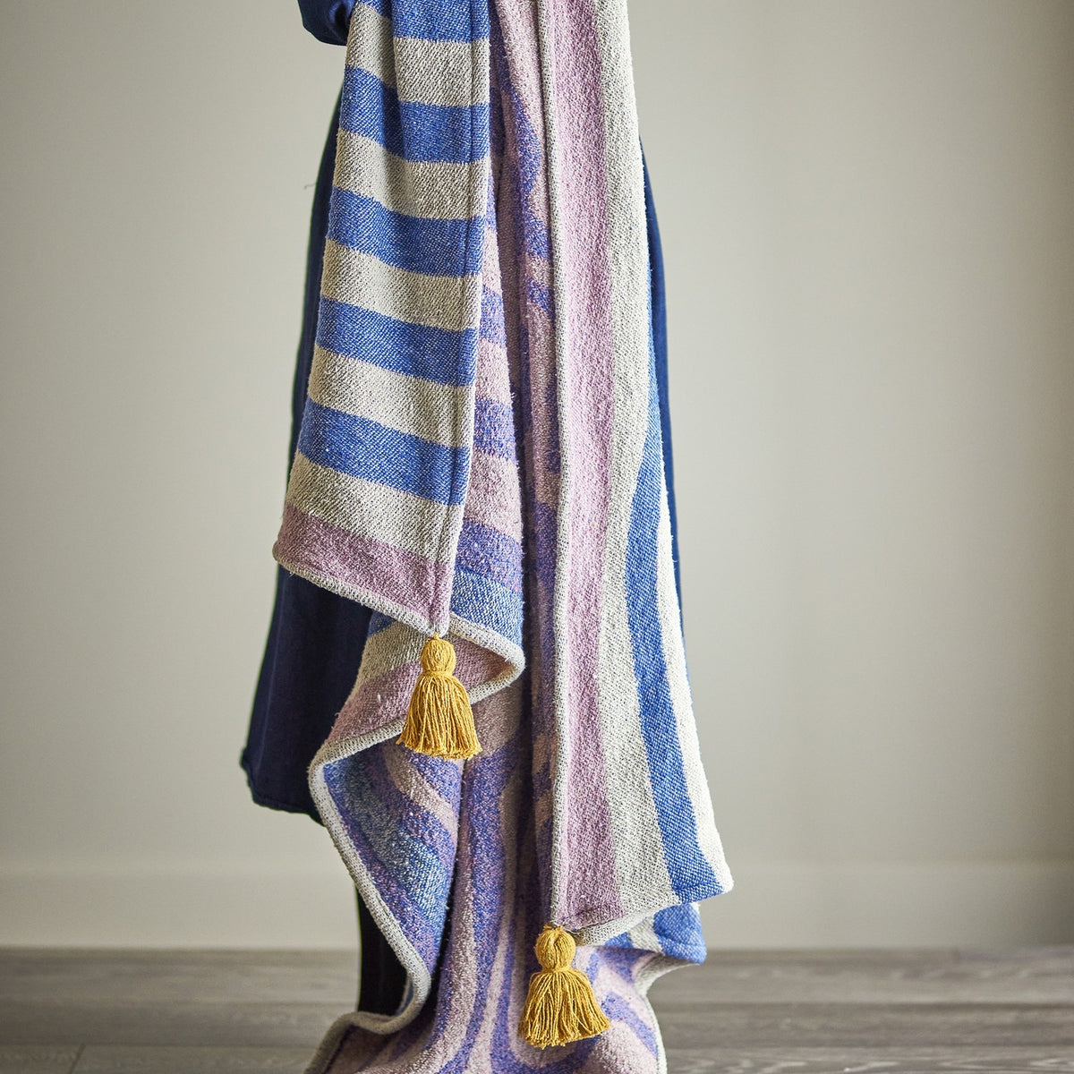 Arlie Throw in blue, a woven fabric throw with striped and swirling patterns and mustard tassel corners, draped over a shoulder against a plain light background.