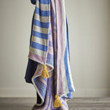Arlie Throw in blue, a woven fabric throw with striped and swirling patterns and mustard tassel corners, draped over a shoulder against a plain light background.