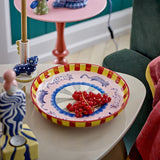 Missy Striped Stoneware Tray in multi colourway, round deep-sided ceramic serving dish with red-yellow striped rim and handpainted cat motif, shown on a side table holding fresh redcurrants.