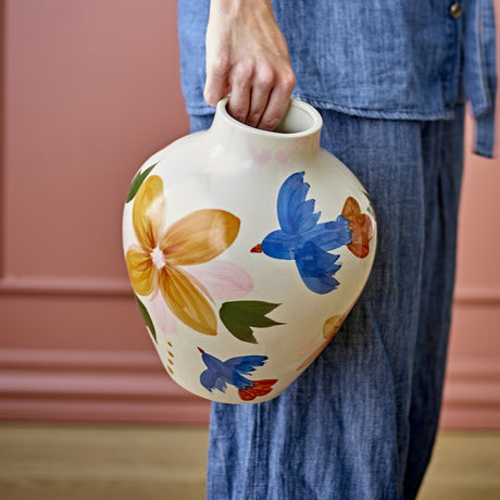 Maricala Floral Stoneware Vase 27cm in white with hand-painted blue birds and yellow flowers, held by the neck against a pink wall.
