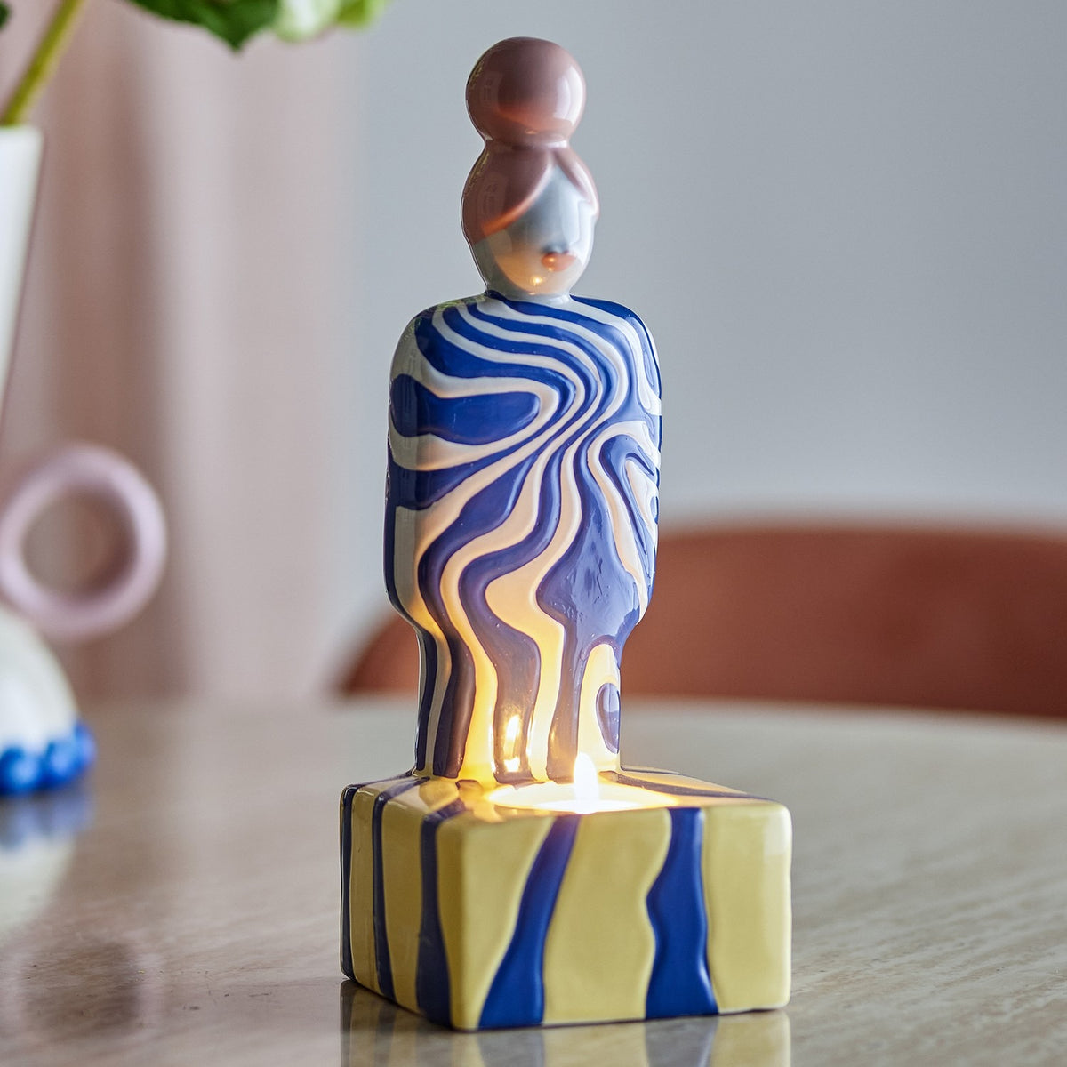 Priya Stoneware Tealight Holder in Multi, a ceramic figure with a bun hairstyle, blue and white swirl-patterned body, and a yellow striped block base holding a lit tealight, shown on a table.