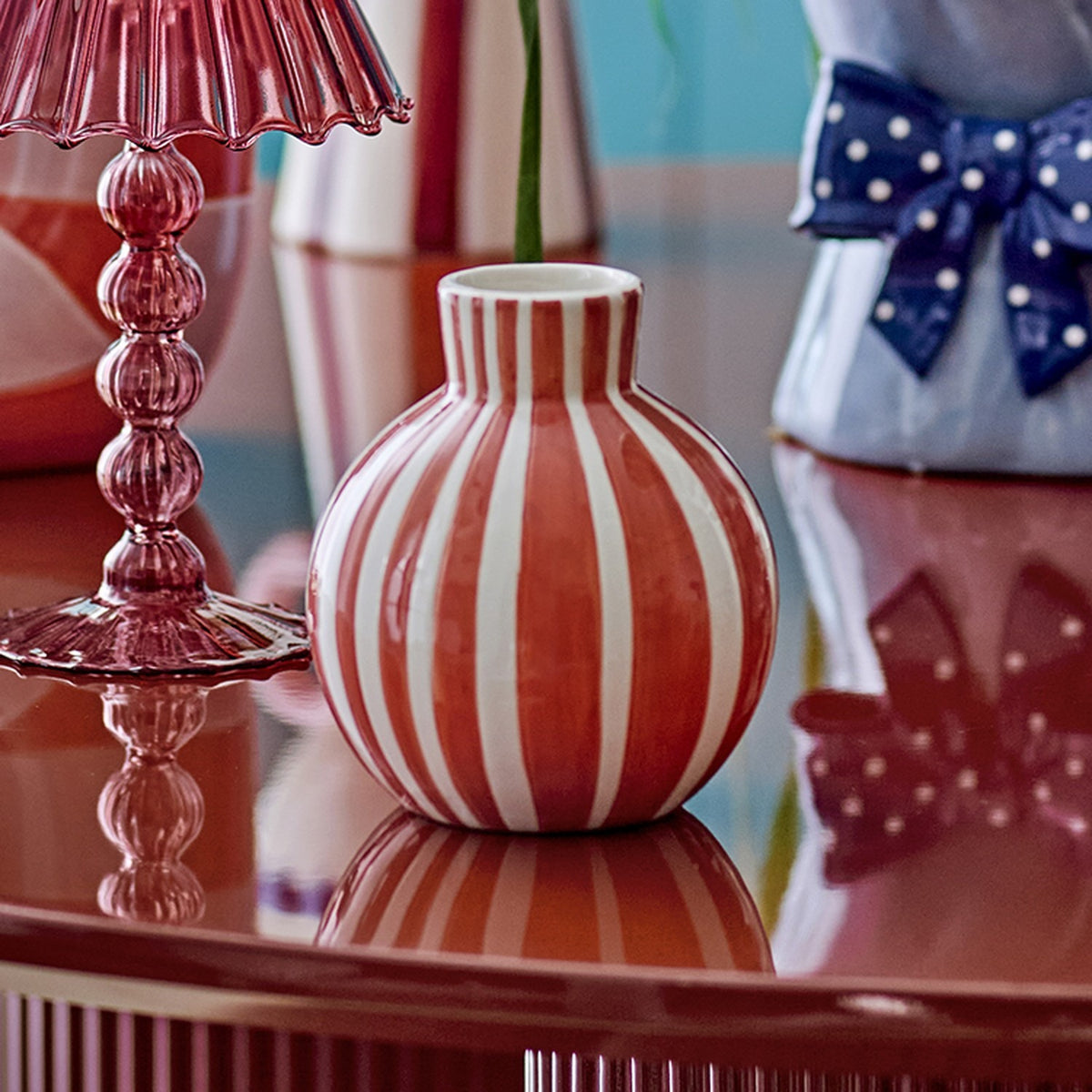 Fian Striped Ceramic Vase 11cm in orange, round hand-painted dolomite vase with a short neck, shown on a reflective table beside a pink glass lamp and polka-dot bow decor.