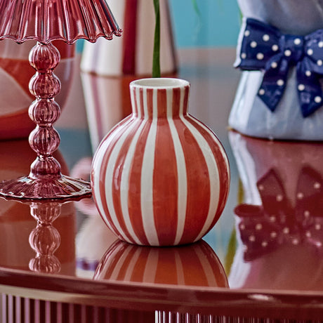 Fian Striped Ceramic Vase 11cm in orange, round hand-painted dolomite vase with a short neck, shown on a reflective table beside a pink glass lamp and polka-dot bow decor.