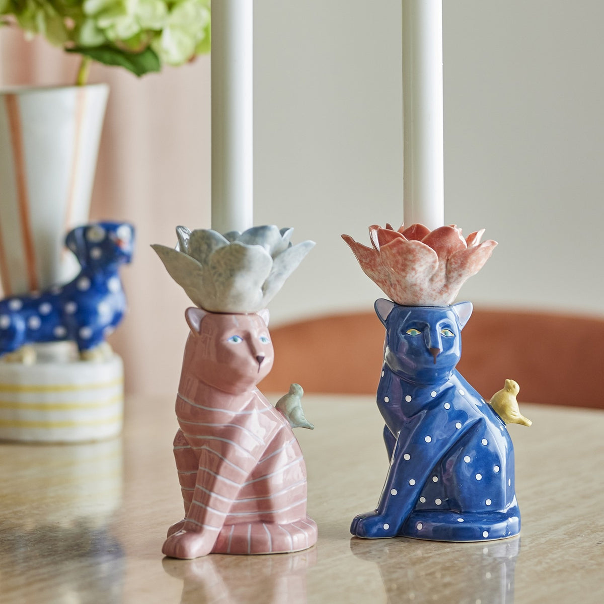 Eri Cat Stoneware Candle Holders set of 2, seated cats in pink stripes and blue polka dots with flower-shaped candle cups and perched birds, shown on a table with white taper candles.