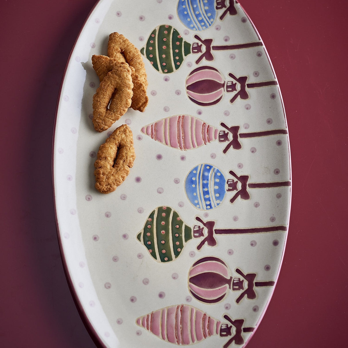 Oval stoneware Christmas platter with hand-painted multicoloured hanging baubles and bows on a dotted ground, shown holding pastries on a dark red surface.