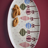 Oval stoneware Christmas platter with hand-painted multicoloured hanging baubles and bows on a dotted ground, shown holding pastries on a dark red surface.