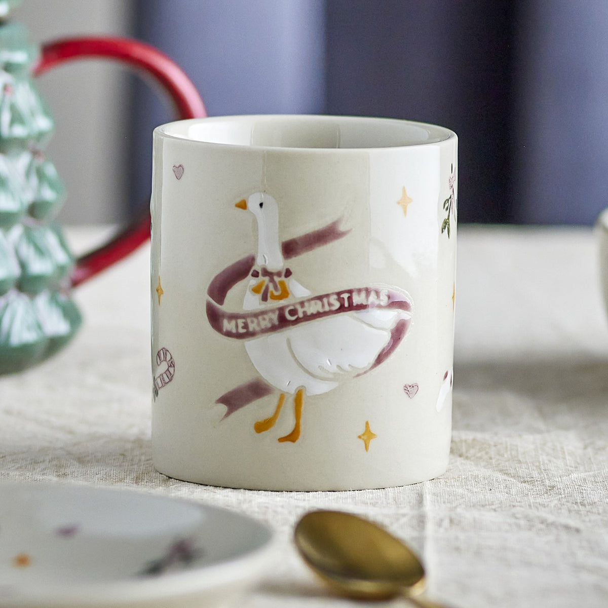 Cream stoneware Christmas mug with embossed goose in a "Merry Christmas" ribbon, surrounded by hand-painted stars, hearts and festive motifs, shown on a table with a teapot and spoon.