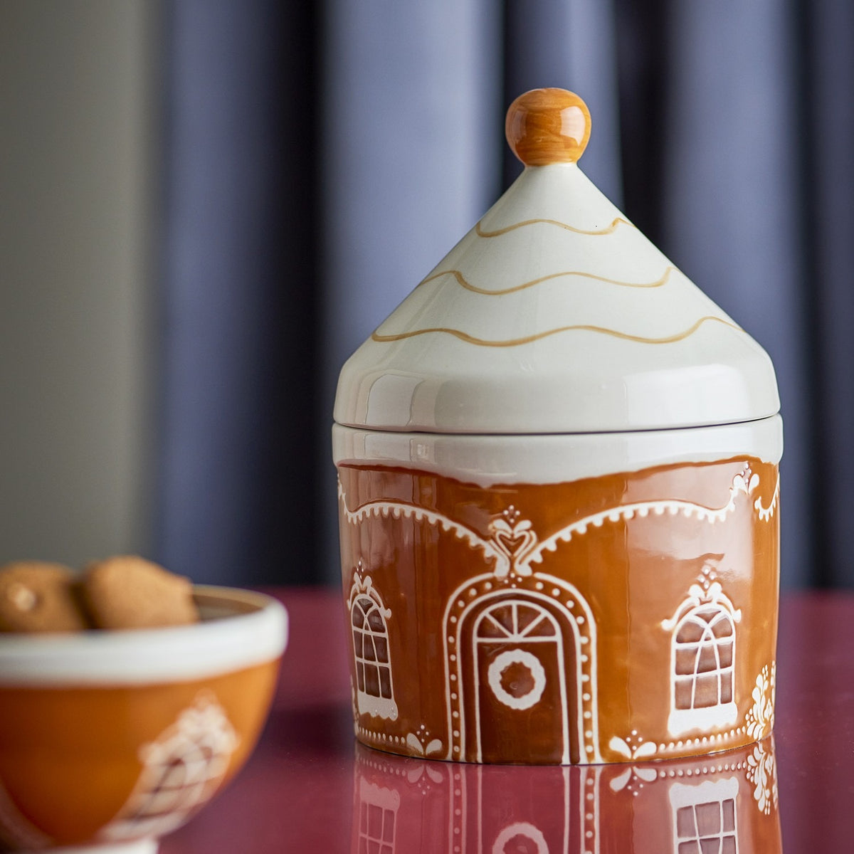 Gina stoneware Christmas storage jar, 1150ml, brown ceramic gingerbread house design with cone-shaped lid and round knob, shown beside a matching bowl of biscuits.