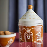 Gina stoneware Christmas storage jar, 1150ml, brown ceramic gingerbread house design with cone-shaped lid and round knob, shown beside a matching bowl of biscuits.