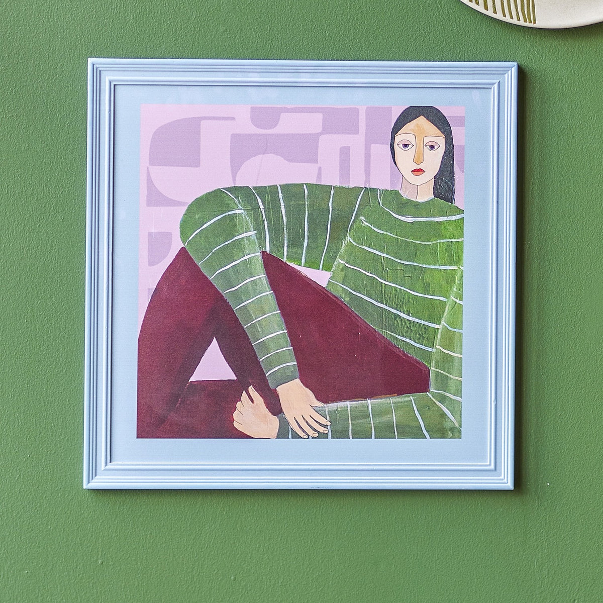 Zoy Wooden Framed Print in Multi, a square wall art piece in a light blue frame featuring a seated woman in a striped green jumper and maroon trousers on a pink abstract background.