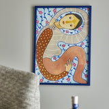 Floriesta Floral Wooden Framed Print in Multi, depicting a hooded figure curled in an S-shape amid floral vines, shown in a blue wooden frame on a living room wall.
