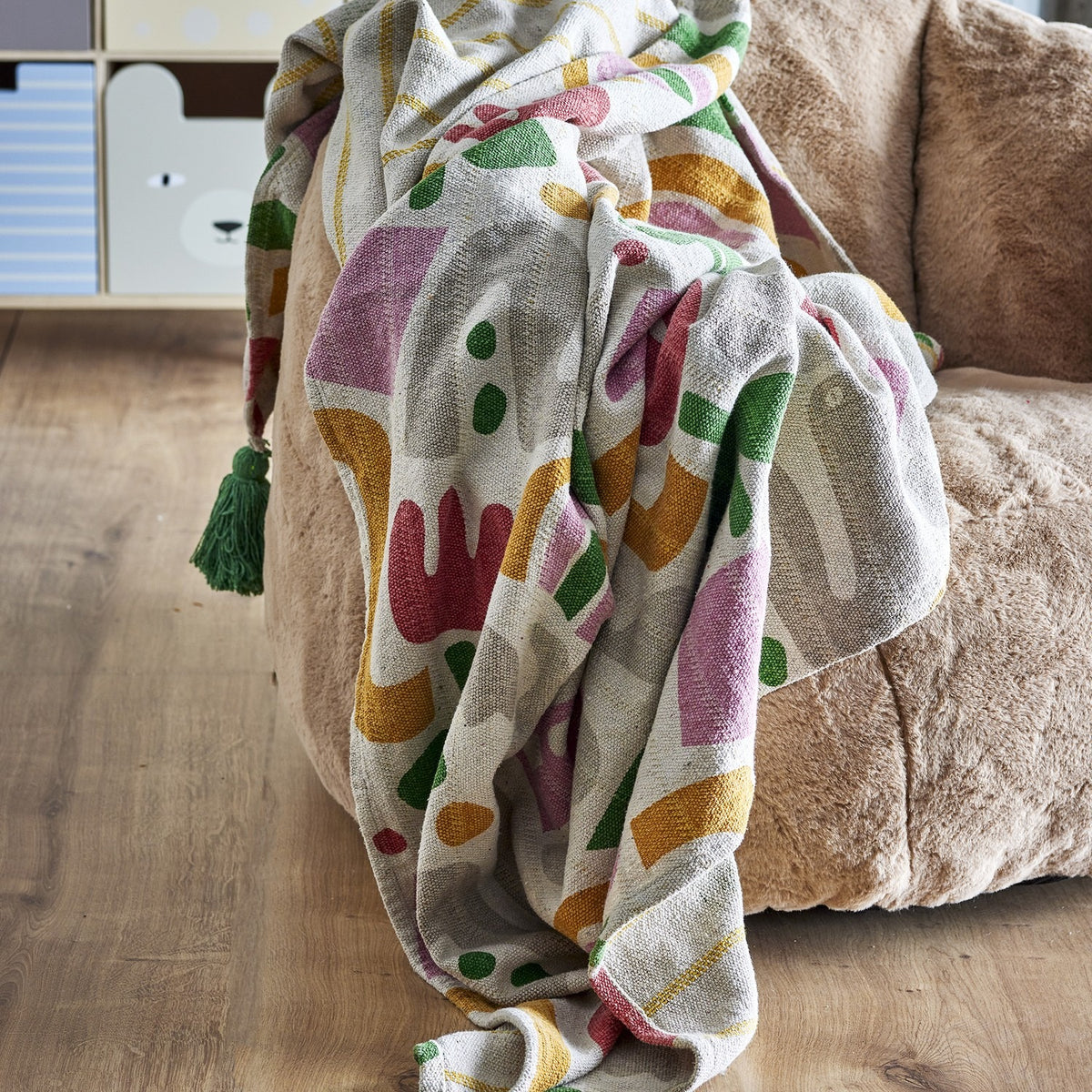 Filur Cotton Throw in multicoloured abstract print with a green tassel corner, draped over a beige upholstered armchair in a children's room setting.