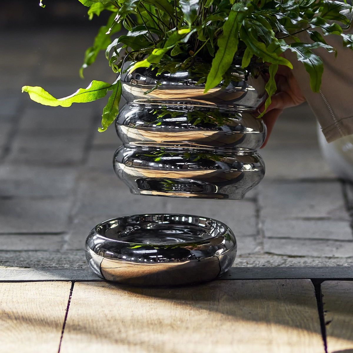 Shine Stoneware Plant Pot with Saucer in silver, its stacked rounded rings reflecting light, shown with a leafy fern above the matching round saucer on a stone and wood surface.