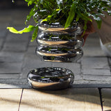 Shine Stoneware Plant Pot with Saucer in silver, its stacked rounded rings reflecting light, shown with a leafy fern above the matching round saucer on a stone and wood surface.