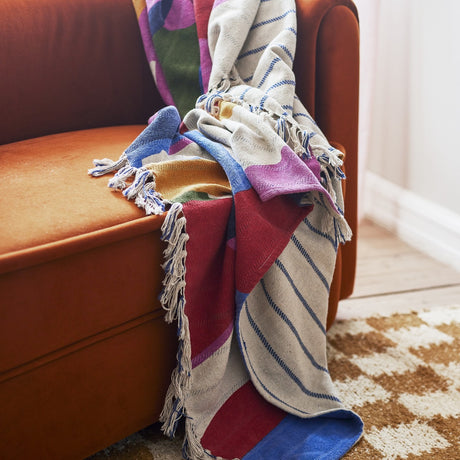 Artista Cotton Throw in multicoloured abstract print with blue striped panel and knotted tassel fringe, draped over a burnt orange armchair.