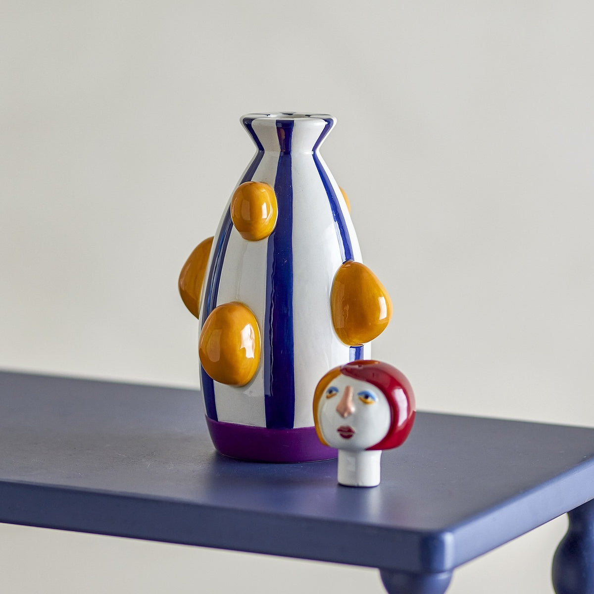 Conneli Ceramic Candle Holder in multicoloured dolomite, tall striped bottle-shaped body with raised bumps, shown with its painted face and bobbed-hair head removed on a blue table.
