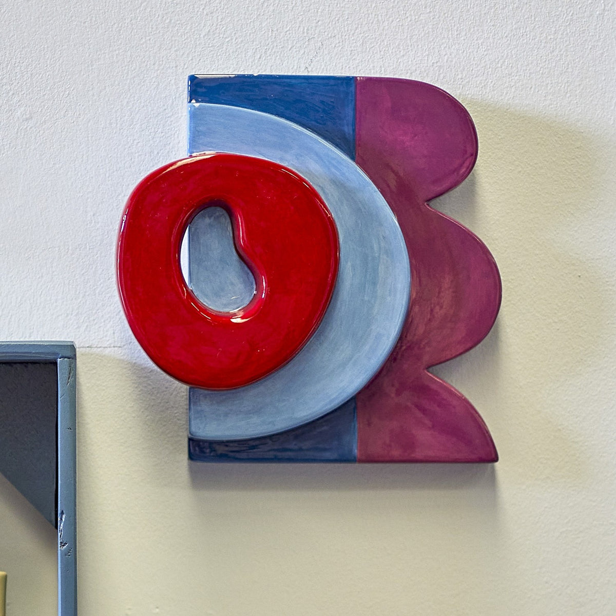 Polivia Ceramic Wall Decor in Multi, a handpainted dolomite piece with a glossy red ring over a pale blue arc, navy and plum panels, mounted on a light wall.