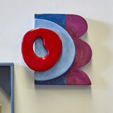 Polivia Ceramic Wall Decor in Multi, a handpainted dolomite piece with a glossy red ring over a pale blue arc, navy and plum panels, mounted on a light wall.