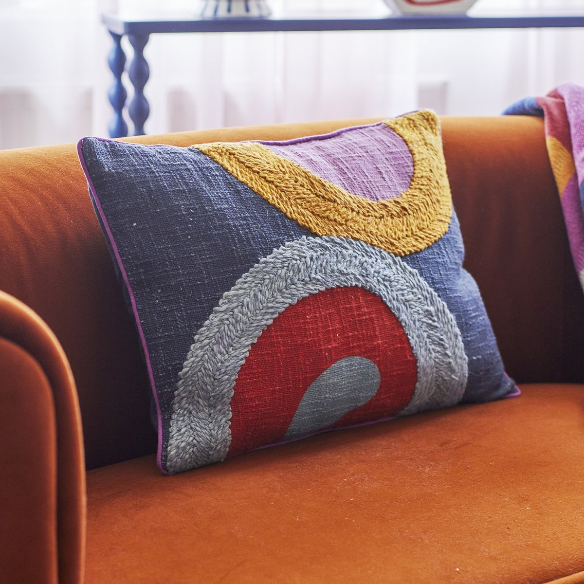 Polivia Cotton Cushion in Multi, rectangular with embroidered curved bands in mustard, lilac, red, and grey on a navy cotton ground, shown resting on an orange velvet sofa.