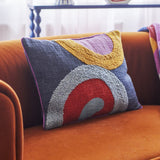 Polivia Cotton Cushion in Multi, rectangular with embroidered curved bands in mustard, lilac, red, and grey on a navy cotton ground, shown resting on an orange velvet sofa.