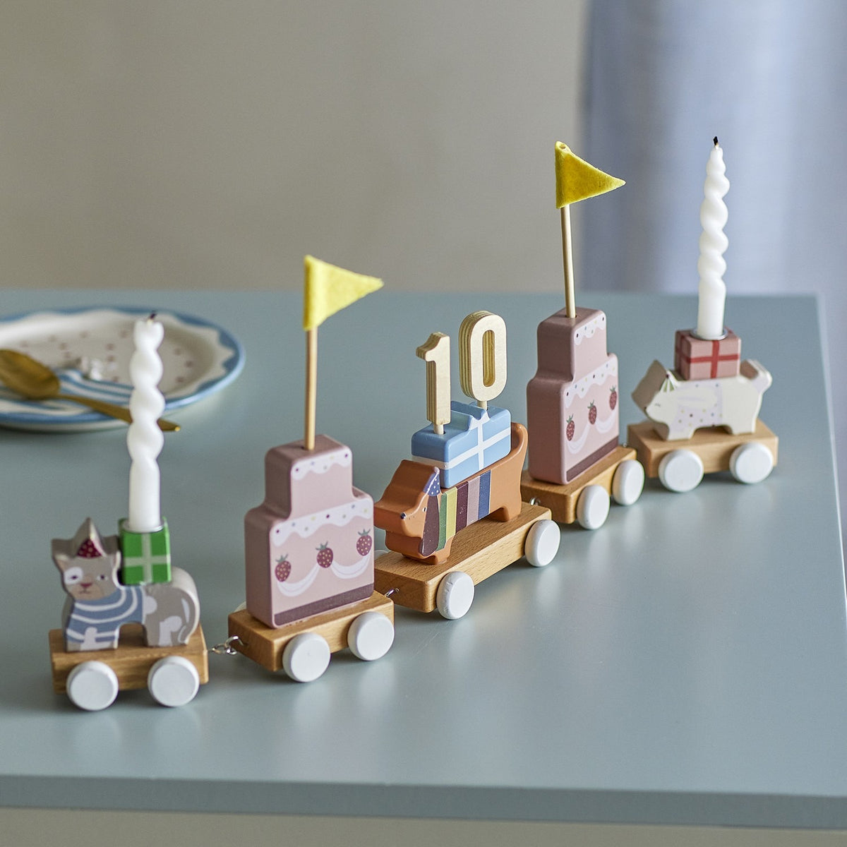 Wooden birthday train with five linked wagons featuring a cat, tiered cakes with yellow pennant flags, a dachshund carrying a 10 topper, and a sheep with a spiral candle, on a grey tabletop.