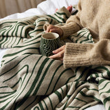 Woodsby Striped Cotton Throw in green and cream wavy stripes, draped over a bed with a person in a sweater holding a mug.
