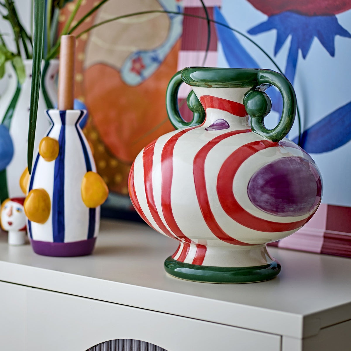 Betiva Striped Stoneware Vase 25.5cm in white and red with a purple spot motif and green rim handles, shown on a sideboard with a smaller striped vase and patterned wall art.
