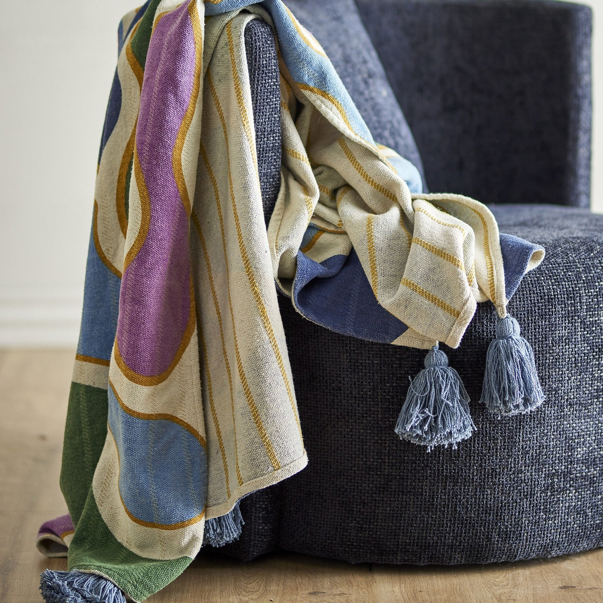 Salacia Throw in Multi draped over a dark upholstered chair, showing curved multicoloured bands, striped sections, and blue tasselled corners.