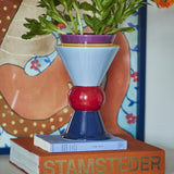 Trilla Ceramic Vase 26.5cm in Multi, a stacked ceramic form with purple rim, pale blue cone, red ball, and navy flared base, holding green foliage on a book atop a decorative scene.