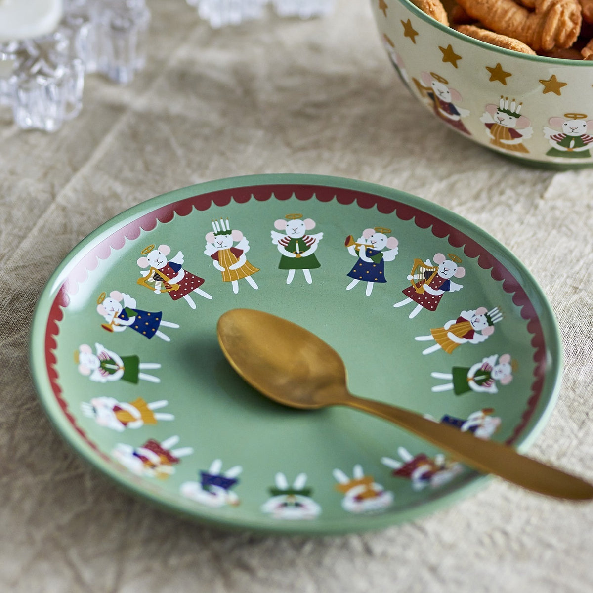 Peo stoneware Christmas side plate in green, featuring a ring of winged angel figures within a scalloped red-edged border, shown with a gold spoon on a linen tablecloth.