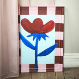 Vinia Floral Wooden Framed Print in Multi, featuring a hand-painted red and blue flower on a pale blue background, set in a pink and brown checked wooden frame, shown leaning against a wall.