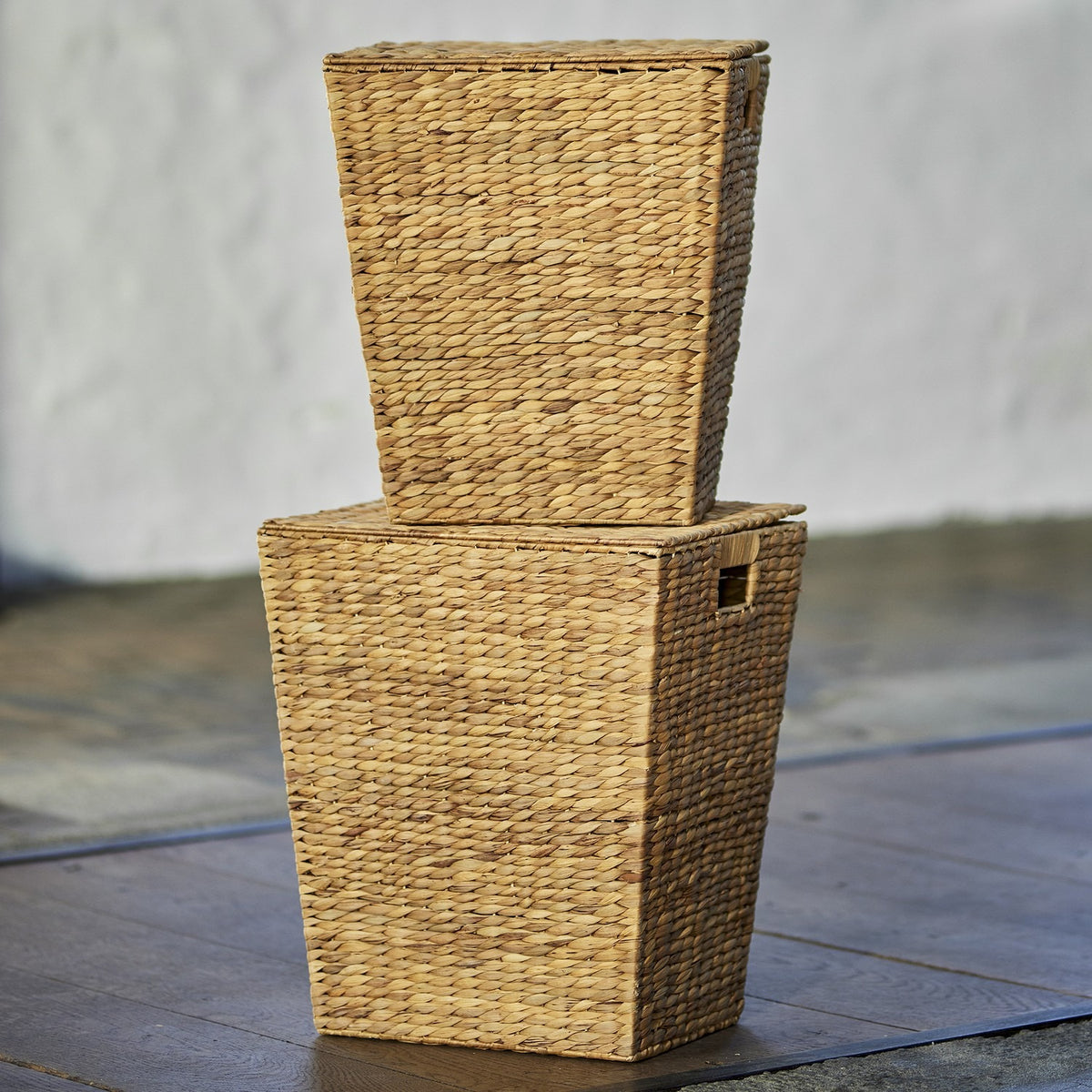 Mios Water Hyacinth Laundry Baskets Set of 2 in natural woven finish, tapered shapes with hinged lids and cut-out handles, stacked outdoors on a wooden deck.