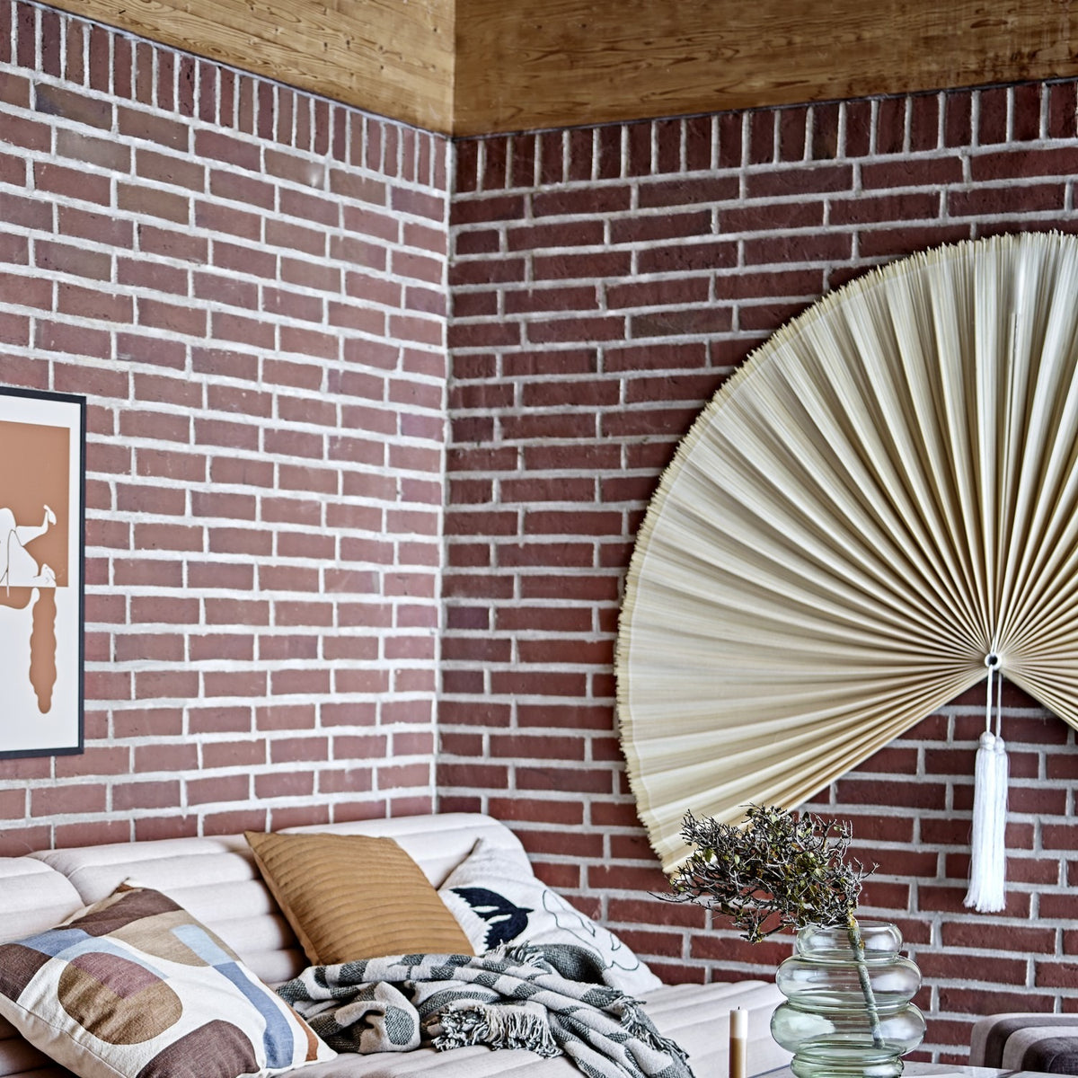 Jaime Bamboo Wall Decor 110cm in natural finish, a large pleated fan-shaped wall art piece with centre tassel, mounted above a living room sofa against a brick wall.