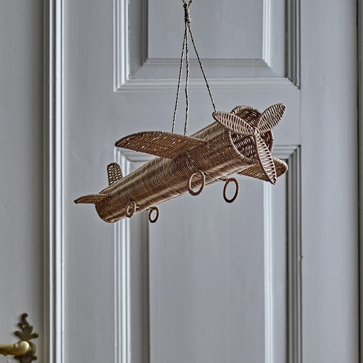 Balazar Rattan Baby Mobile in natural woven rattan, shaped as an aeroplane with wings, tail fin, propeller and wheel-like hoops, hanging by cord against a panelled door.