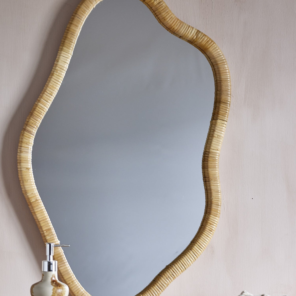 Ashlie Rattan Wall Mirror 50x80cm in natural rattan with a wavy, freeform frame, mounted on a light wall above a ceramic soap dispenser.