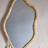 Ashlie Rattan Wall Mirror 50x80cm in natural rattan with a wavy, freeform frame, mounted on a light wall above a ceramic soap dispenser.