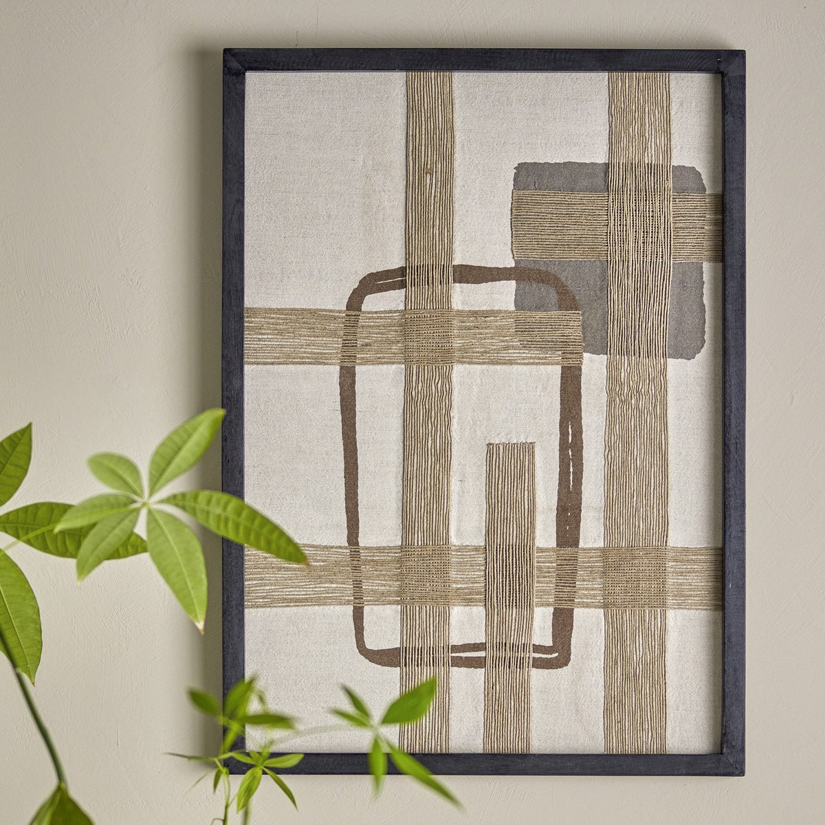 Bie Geometric Cotton Wall Decor in beige, framed textile art with woven jute bands forming a grid over a rounded rectangle outline and square block, shown on a wall beside green foliage.