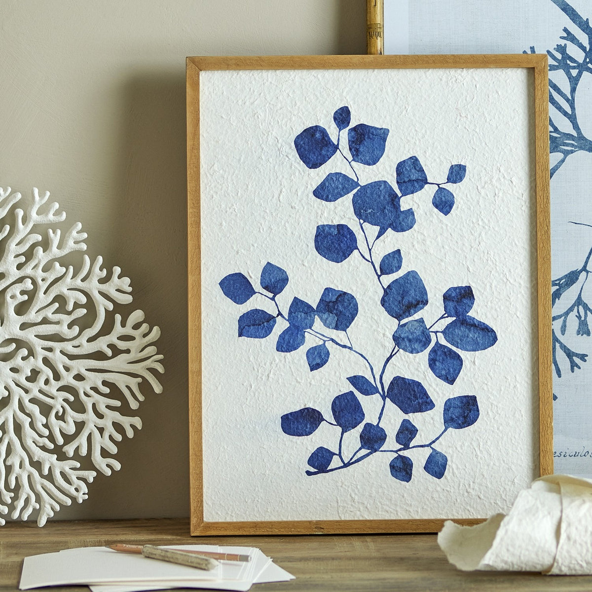 Gunnvor Wooden Wall Decor - Natural wood frame botanical print with a blue leafy branch on textured paper, displayed beside coral decor on a shelf.