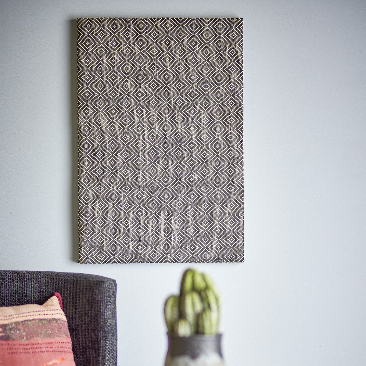 Rivalta Pin Board in black with woven diamond-pattern texture, rectangular MDF frame, shown mounted on a light grey wall above a sofa and cactus planter.