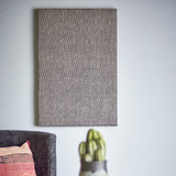 Rivalta Pin Board in black with woven diamond-pattern texture, rectangular MDF frame, shown mounted on a light grey wall above a sofa and cactus planter.