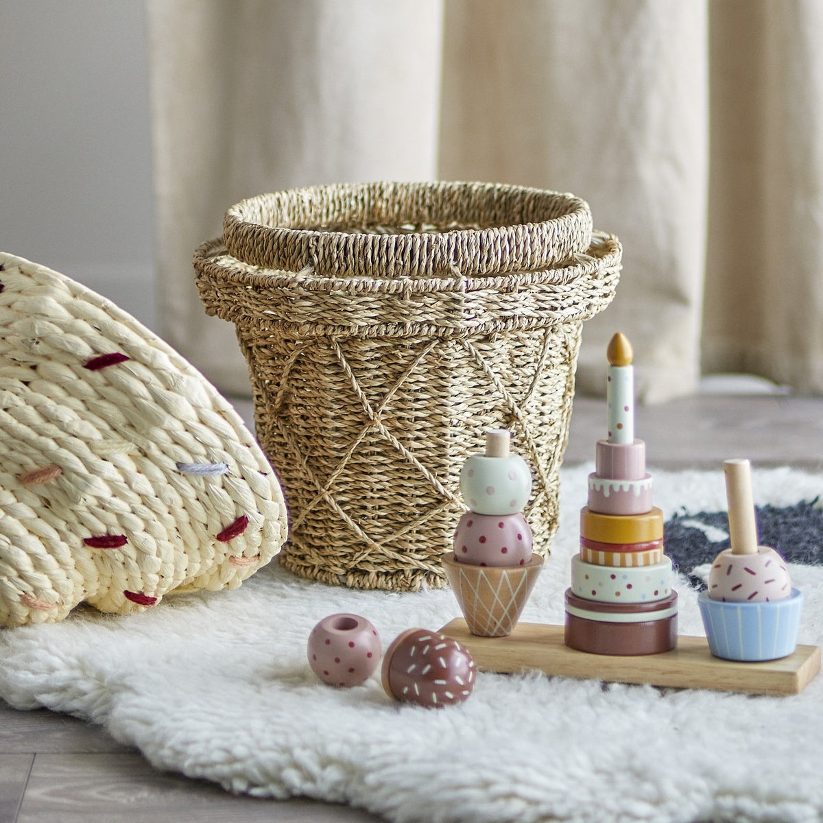 Cillie Seagrass Basket with Lid 41cm in natural, woven ice-cream cone shape with criss-cross lattice base, shown beside stacking toy pieces on a sheepskin rug.
